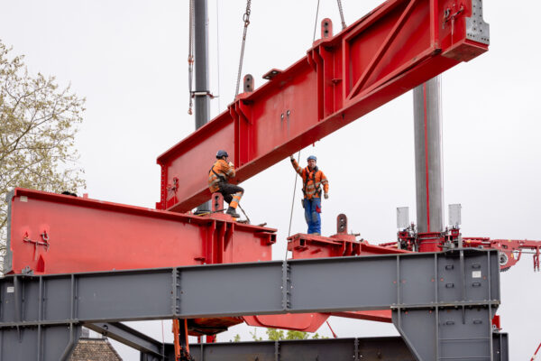 Zwei Bauarbeiter montieren einen Stahlträger einer Portalkonstruktion unter dem angehobenen Kranbauteil.