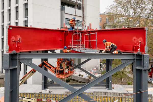 Mehrere Bauarbeiter montieren eine Portalkonstruktion aus Stahlträgern unter Einsatz eines Pneukrans.